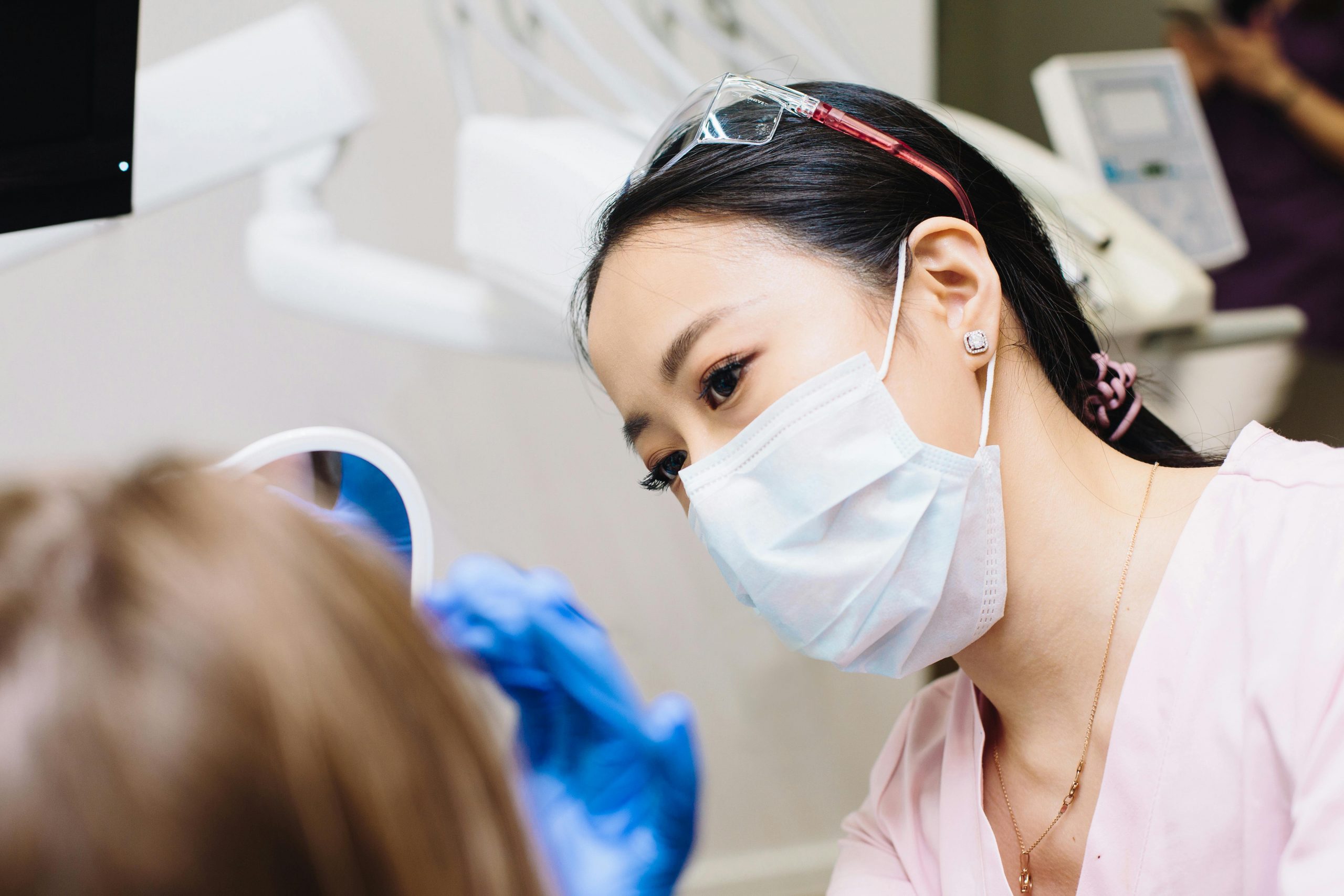 female Asian dentist wearing blue gloves and a mask, examining a patient's mouth